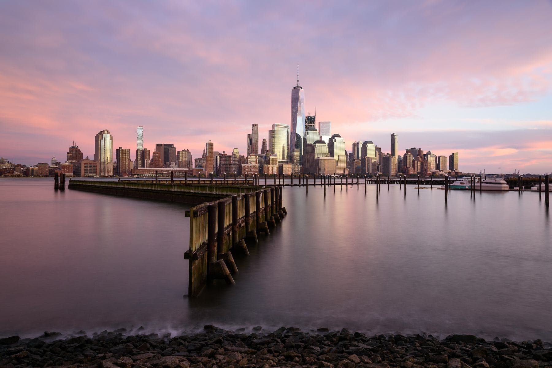 The Lower Manhattan skyline at dusk under a pink sky, seen across the Hudson River from an old pier in Jersey City