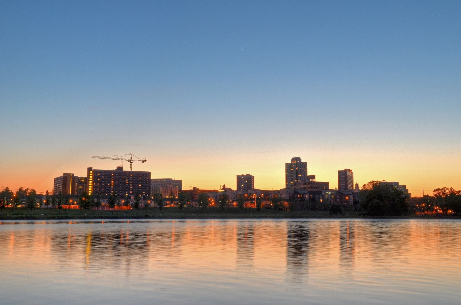 The New Brunswick skyline at sunset, its lit windows reflected in the still Raritan River