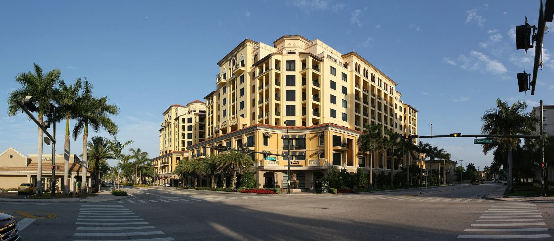 A cream Mediterranean-style building with arched balconies at the palm-lined corner of Palmetto Park Road and Mizner Boulevard under a blue sky