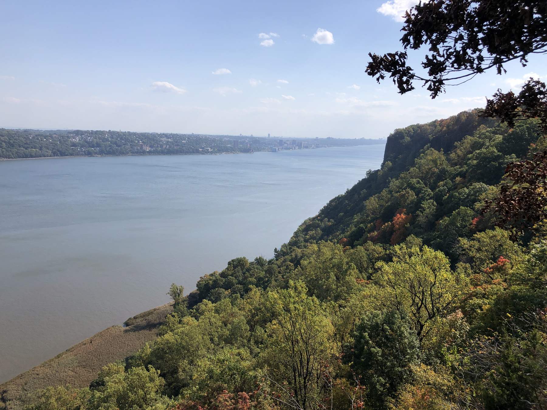 The wide Hudson River seen from the wooded Palisades cliffs in early autumn, with the far shore and a pale blue sky