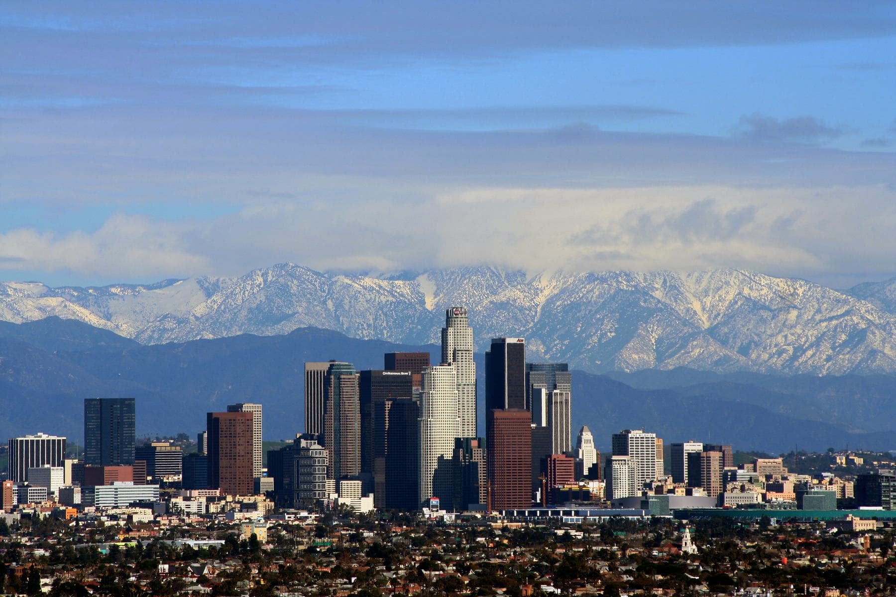Downtown Los Angeles skyscrapers in front of snow-covered San Gabriel Mountains under a streaked blue sky
