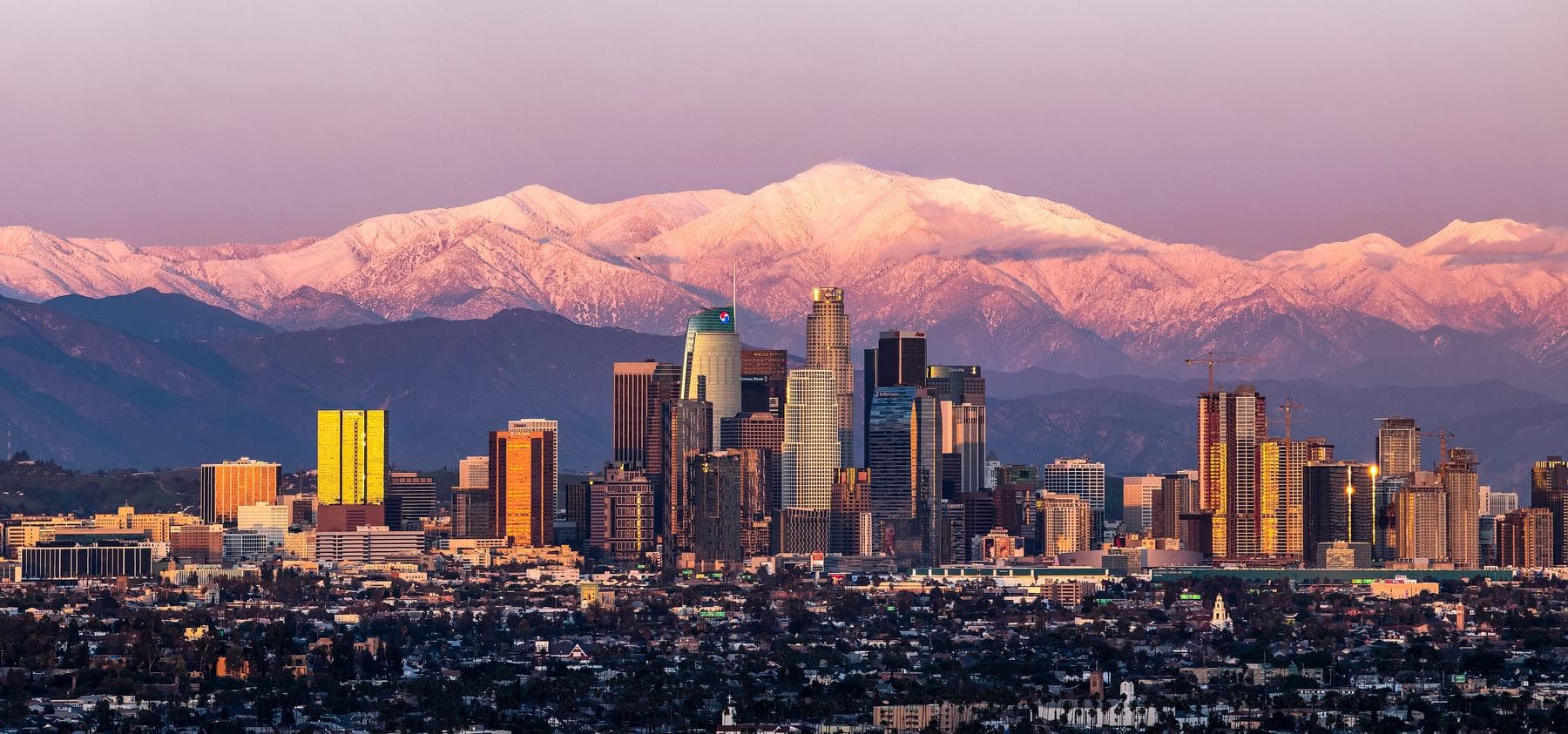 Downtown Los Angeles skyline at sunset, lit gold, with snow-capped mountains glowing pink behind it