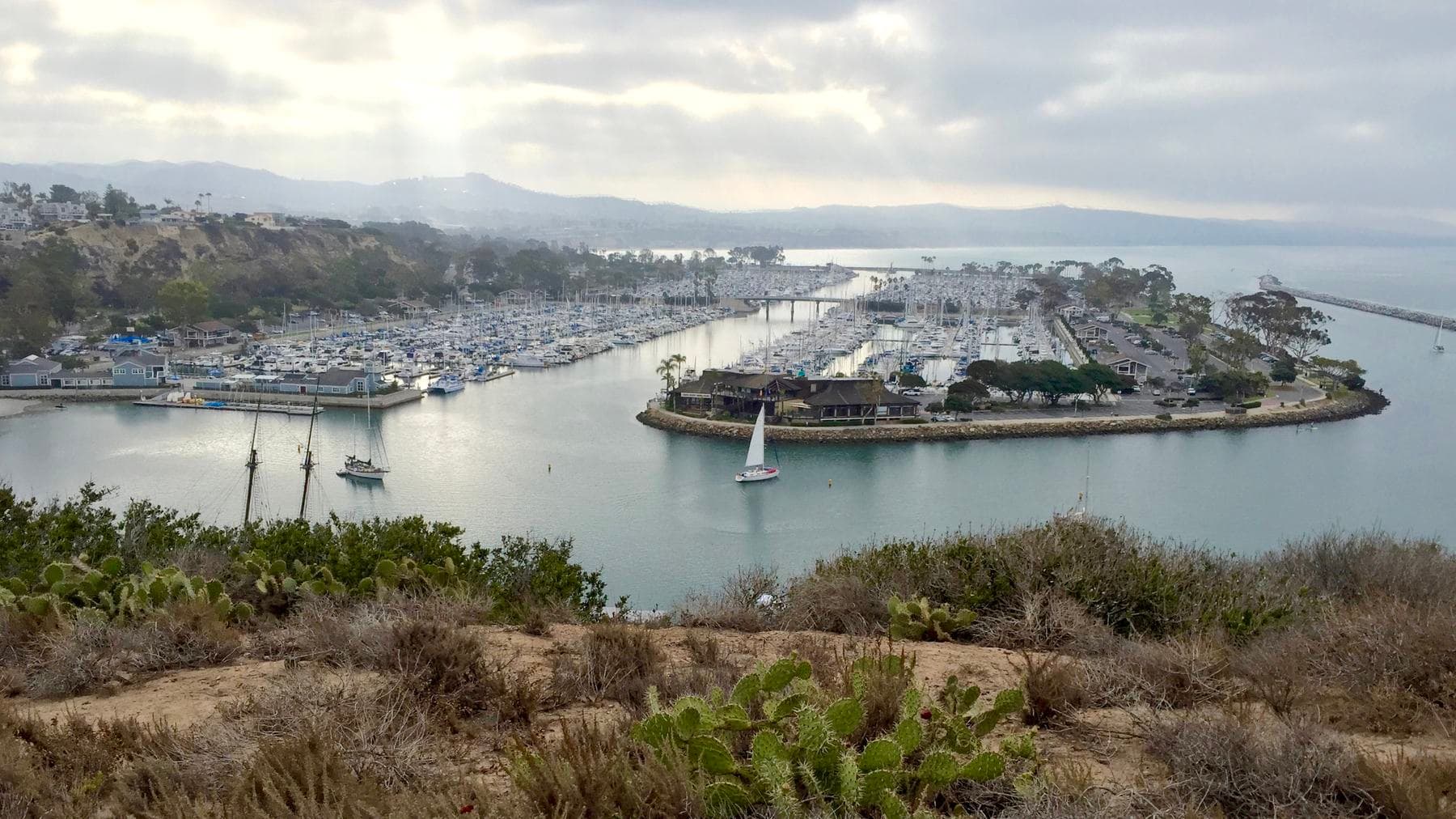 Dana Point Harbor from the headland above, with marinas, a sailboat in the channel and hills under soft cloud