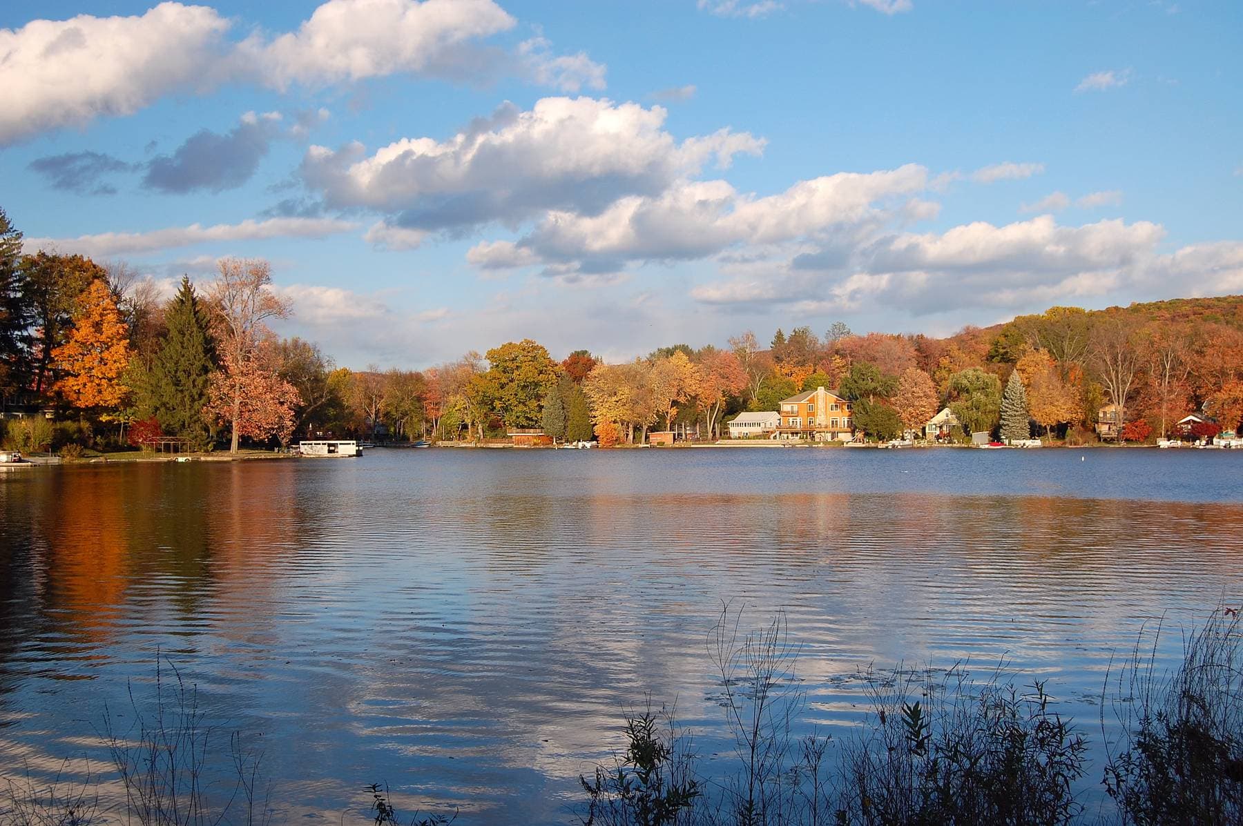Autumn trees and lakeside houses reflected in Greenwood Lake under a blue sky with white clouds