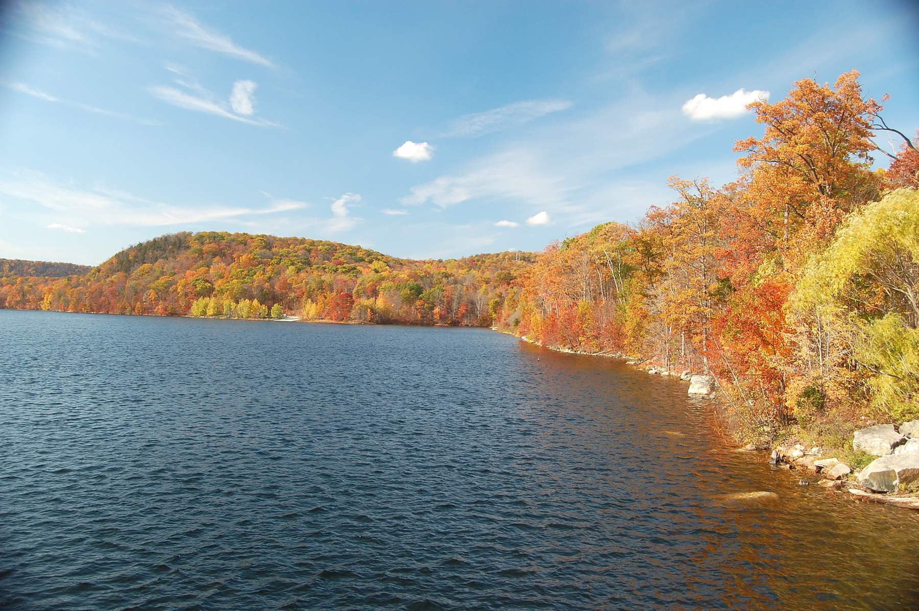 Monksville Reservoir with a hillside of orange and red autumn trees along the shore under a blue sky