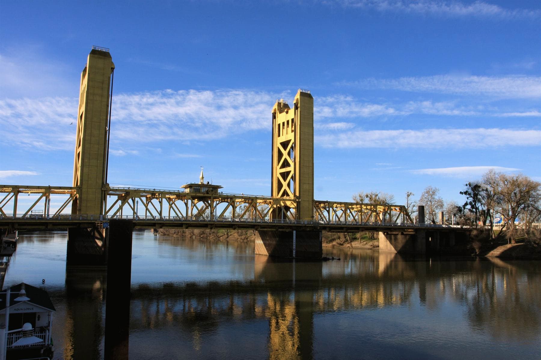 The gold Tower Bridge over the Sacramento River, reflected in calm water under a blue sky with thin clouds